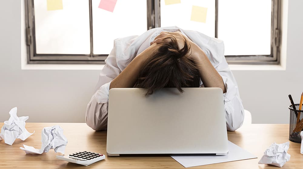 Personne épuisée devant un ordinateur portable, entourée de papiers froissés.