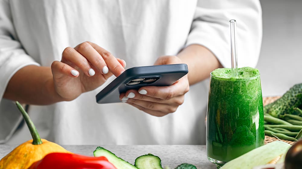 Personne utilisant un smartphone près de légumes et d'un smoothie vert.