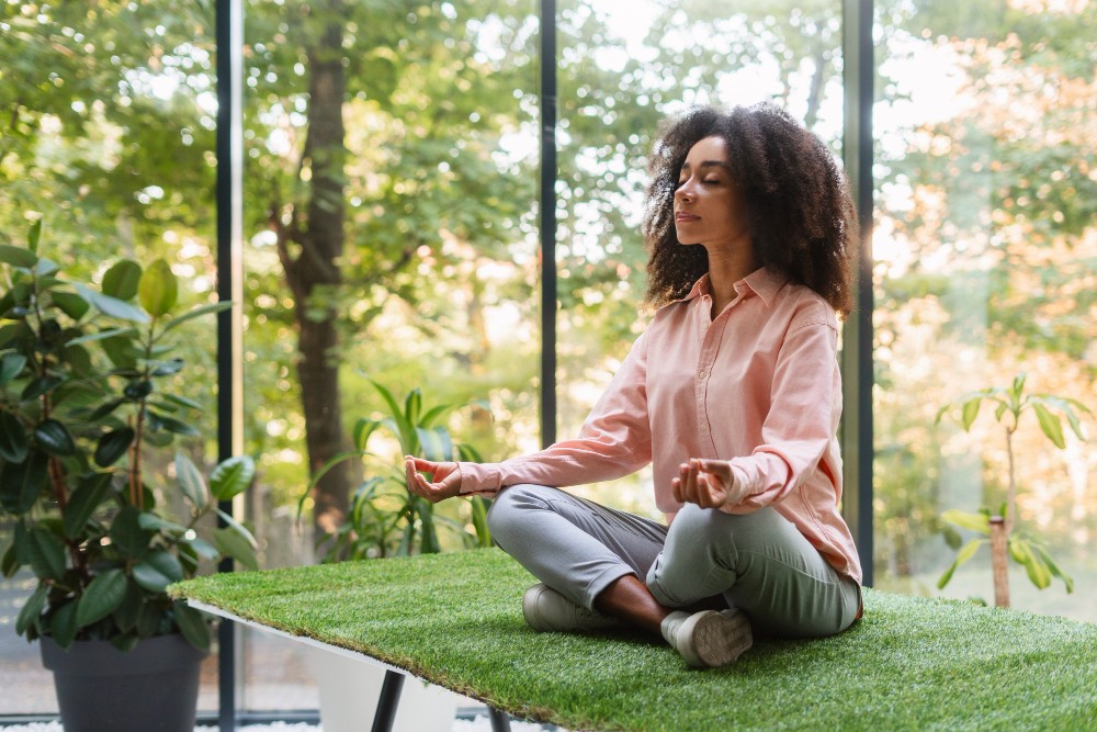 Femme méditant sur une table recouverte de gazon, entourée de plantes.