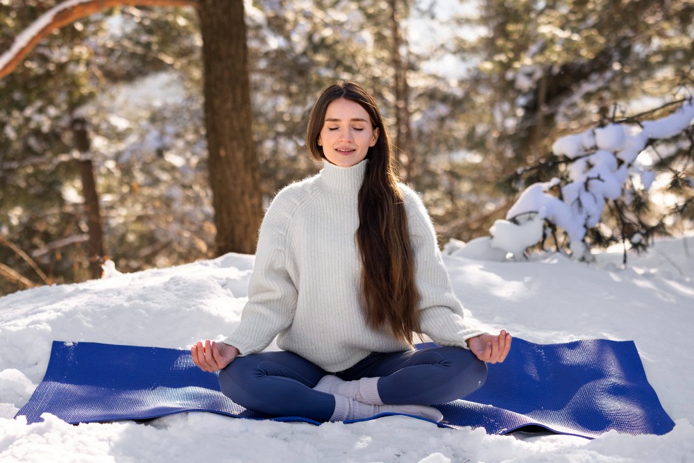 Femme méditant sur un tapis de yoga dans la neige, entourée d'arbres.