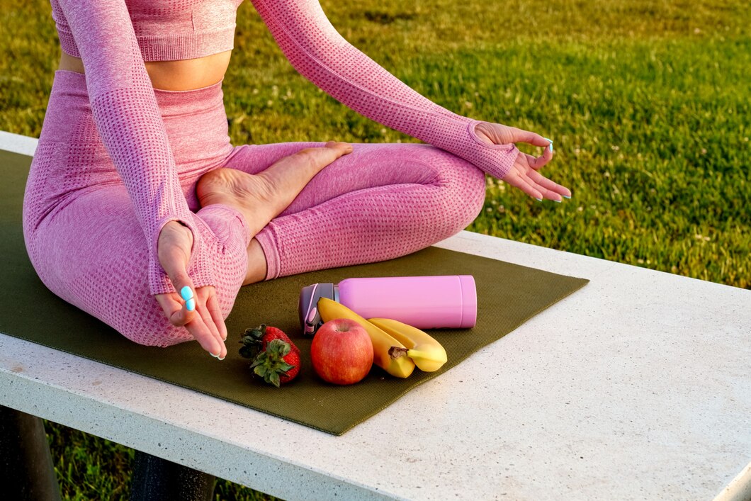 Femme en tenue rose méditant sur un tapis avec fruits et bouteille rose.