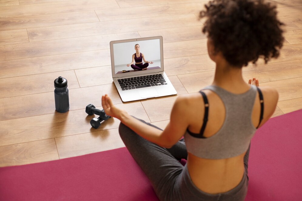 Femme faisant du yoga devant un ordinateur portable, haltères et gourde à côté.